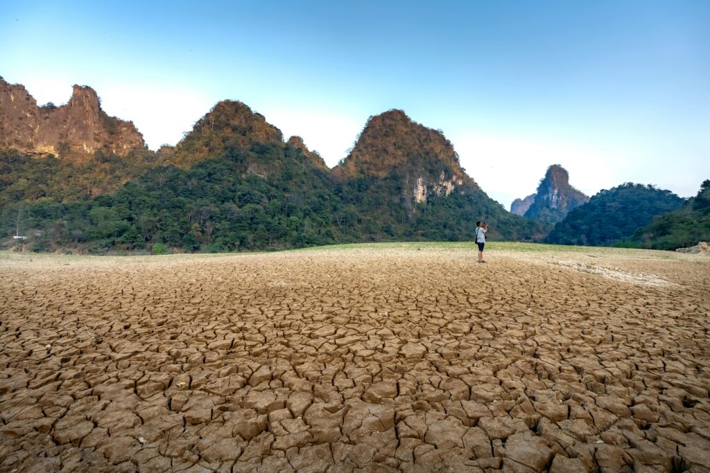 A lone photographer stands on cracked earth with towering mountains in the background under a clear sky.