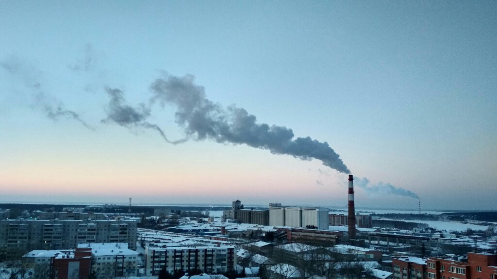 A cityscape featuring industrial smokestacks emitting smoke against a sunset sky, highlighting environmental pollution.